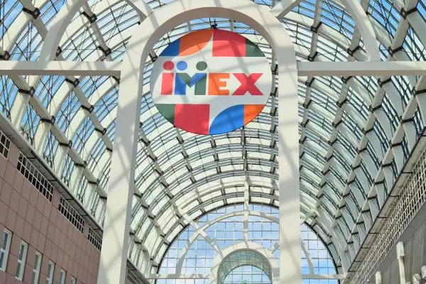 Glasdach einer Messehalle mit großem bunten IMEX-Logo im Zentrum, umgeben von weißen Stahlträgern und blauem Himmel.