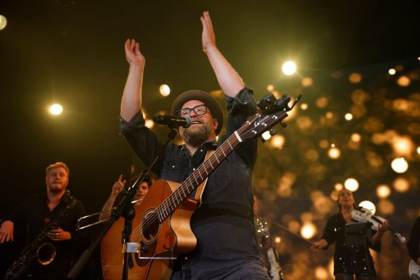 Ein Mann mit Bart, Brille und Hut spielt auf einer Bühne eine Akustikgitarre und singt in ein Mikrofon, wobei er die Arme über seinen Kopf gestreckt hat. Im Hintergrund sind weitere Musiker und eine helle, unscharfe Beleuchtung zu sehen.