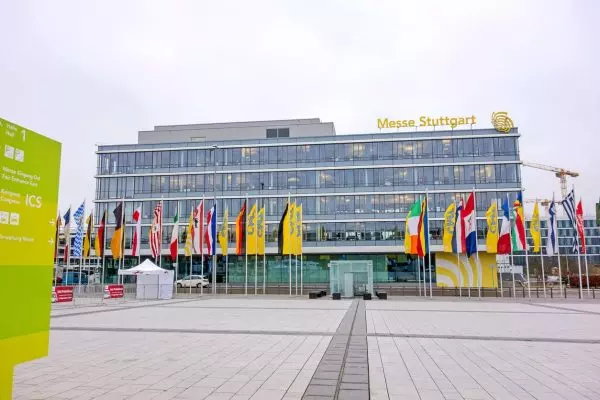 stuttgart Außenansicht des Verwaltungsgebäudes der Messe Stuttgart bei bewölktem Himmel. Im Vordergrund befindet sich ein gepflasterter Vorplatz mit einer langen Reihe internationaler Flaggen und Fahnen. Das moderne Gebäude verfügt über eine Glasfassade und trägt den gelben Schriftzug „Messe Stuttgart“ auf dem Dach. Links im Bild ist eine grüne Hinweistafel für den Eingang Ost und das ICS (International Congress Center Stuttgart) zu sehen.