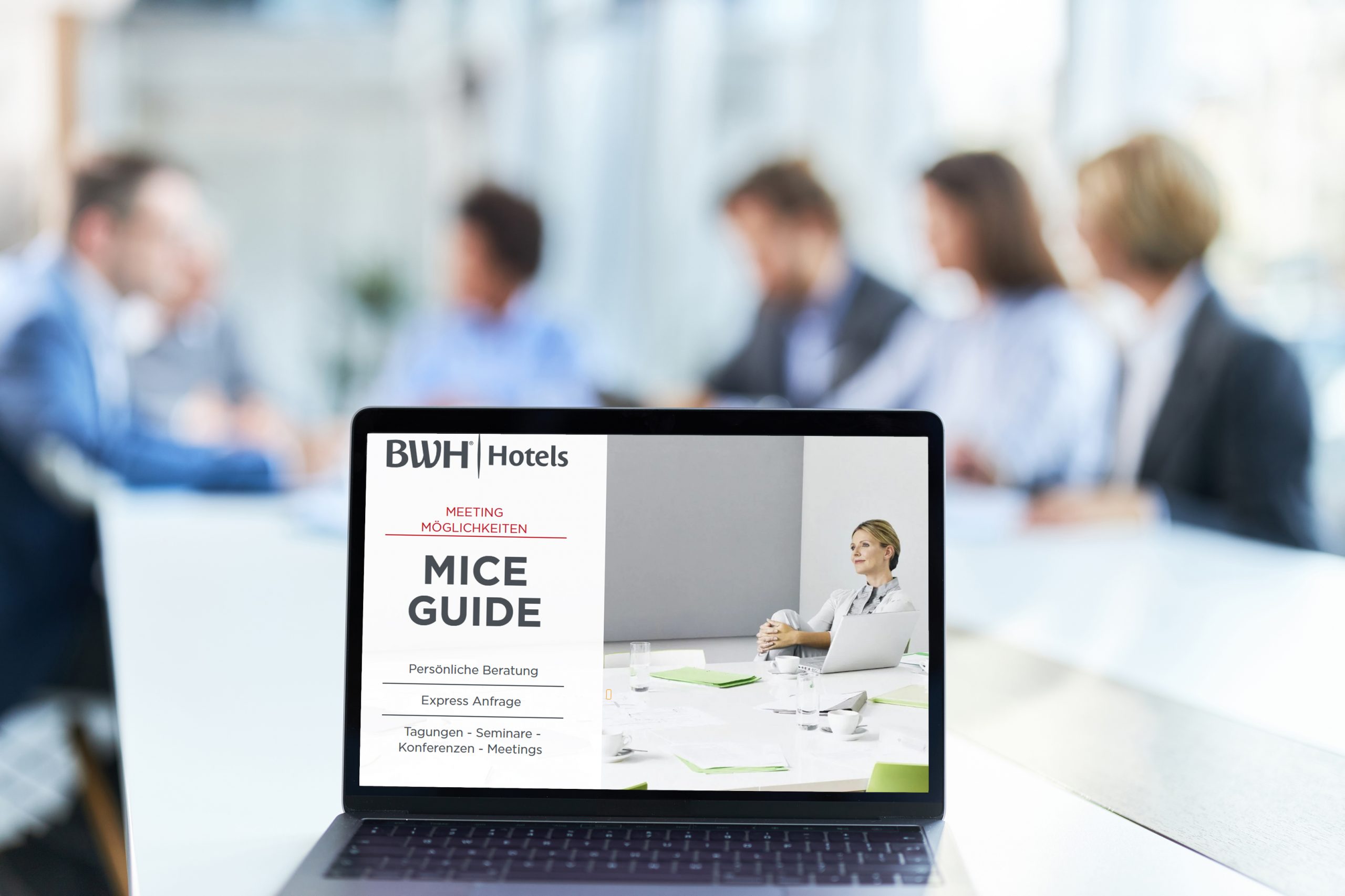 Blank white screen of a computer on a desk in the office. Blick auf einen Computer. Im Hintergrund Personengruppe im Gespräch