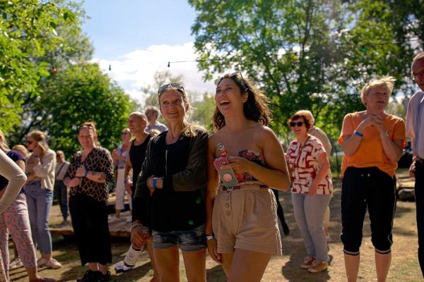 Eine Gruppe fröhlicher Menschen bei einem Outdoor-Event im Grünen; zwei lachende Frauen stehen im Zentrum des sonnigen Geschehens.