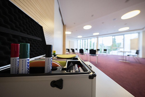 Im Vordergrund Moderatorenkoffer im Hintergrund Stuhlkreis und große Fensterfront